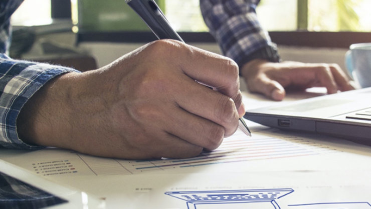 Nahaufnahme einer Hand, die mit einem Stift auf einem Dokument schreibt, neben einem geöffneten Laptop. Auf dem Papier sind farbige Linien. Im Hintergrund unscharfe Fenster mit Tageslicht.