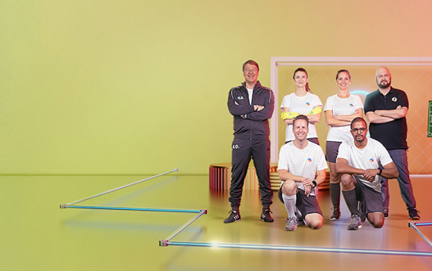 Sechsköpfiges Team posiert vor farbigem Hintergrund mit Taktiktafel und Tor.