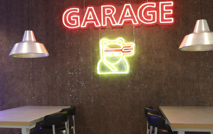Two tables beneath hanging lamps, with neon signs reading "GARAGE", a robot, and a ninja cat on the wall in the background.