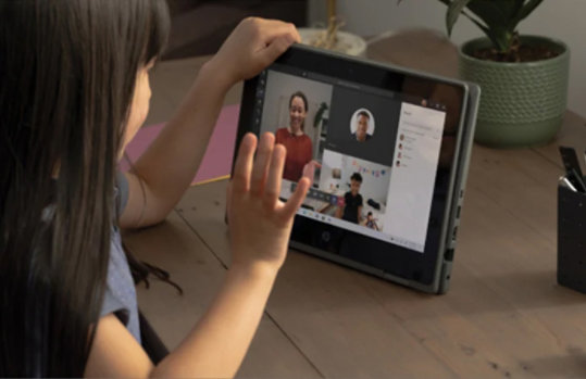 Ein junges Mädchen sitzt zuhause an einem Tisch vor einem Tablet. Sie winkt dabei in die Kamera des Tablets. Auf dem Screen des Tablets sind weitere Personen zu sehen, die sich ebenfalls in dem Gruppen-Telefonat befinden.