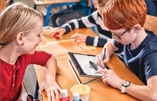 Ein Junge sitzt in einem Klassenzimmer an einem Tisch und malt auf einem Tablet einen Wal, während sich ein Mädchen zu ihm beugt und ebenfalls auf das Tablet schaut.