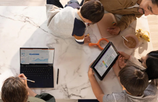 Ein Mann sitzt mit vier Kindern an einem Tisch. Zwei Kinder halten ein Tablet in der Hand und schauen darauf, während ein Junge einen Laptop vor sich hat und parallel etwas auf ein Blatt Papier schreibt.