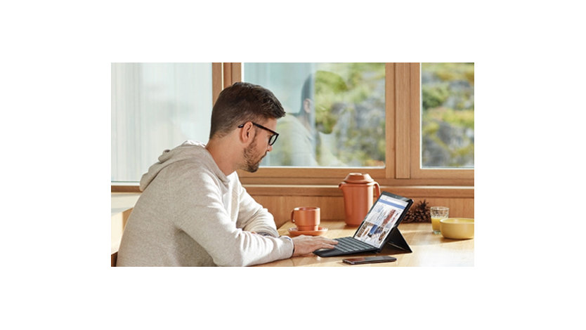 Junger Mann sitzt an einem Schreibtisch mit einer Kaffeetasse und einem Surface Pro 7+ und scrollt durch das Internet. Neben dem Laptop liegt ein Smartphone.