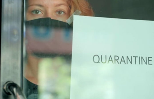 Μια γυναίκα που στέκεται πίσω από μια γυάλινη πόρτα φορώντας μια μάσκα. Στη γυάλινη πόρτα υπάρχει ένα κομμάτι χαρτί στο οποίο είναι γραμμένη η λέξη «Καραντίνα».