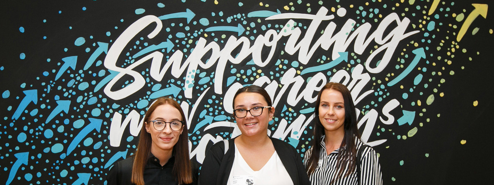 Womens are standing and smiling front of the wall banner | Supporting new career pathways