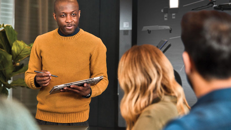 Une personne vêtue d’un chandail jaune, tenant une tablette et un stylo, a engagé une discussion avec deux autres personnes dans un cadre intérieur moderne.