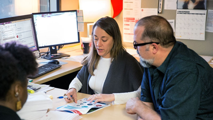 Trois personnes assises autour d’un bureau dans un bureau sont occupées à examiner un document à l’aide de tableaux et de graphiques.