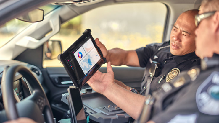 Two police officers sitting in a police car while looking at a Surface Go 3 in a ruggedized case with Teeams screen shown.