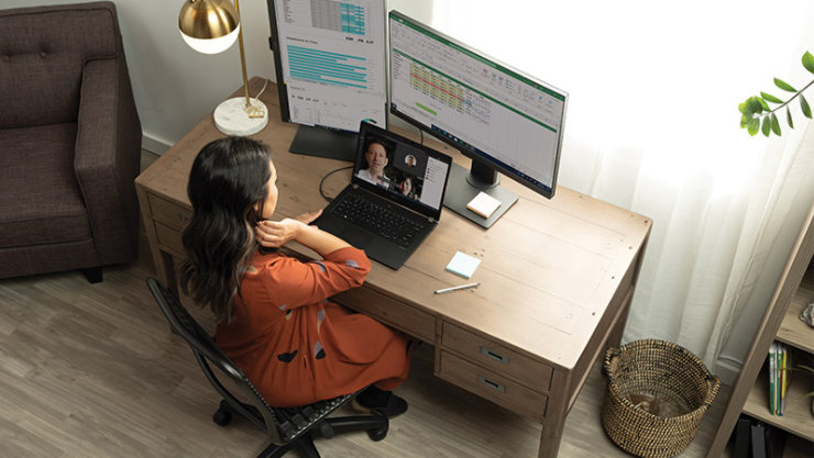 A woman having a Microsoft Teams in her living room home office on her Acer TravelMate P6. The laptop is configured with a multi-monitor display system showing Excel spreadsheets and a PowerBI dashboard. Remote Working collection.