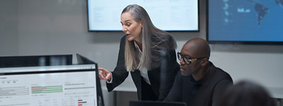 Kobieta gestykuluje w kierunku ekranu komputera, podczas gdy mężczyzna pracuje na innym komputerze. Kilka monitorów z danymi widocznych w tle.