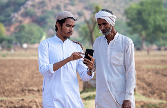 Two farmers standing in an arid village field looking at a mobile phone