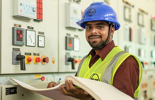 A man wearing a blue hard hat working at a machine while holding a large log book