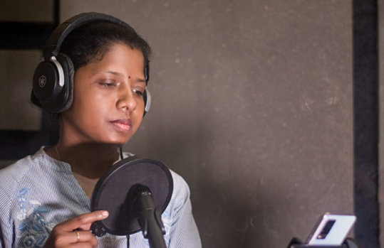 A woman reading aloud text from a smartphone’s display in front of a microphone at a recording studio