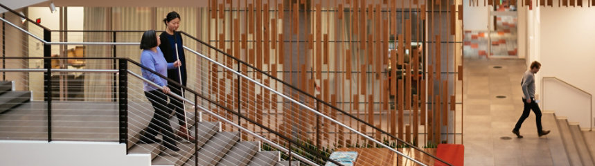 A woman with visual impairment aided by a walking stick taking the stairs while talking to a colleague inside an office.