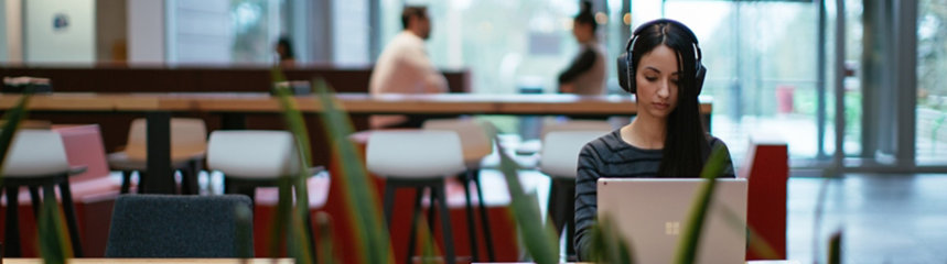 A woman with a cognitive disability, wears headphones and works on a laptop in the café of an office building.