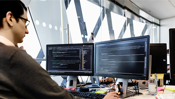Male developer coding in front of two monitors at desk in office. Programming code shown on both monitors. 