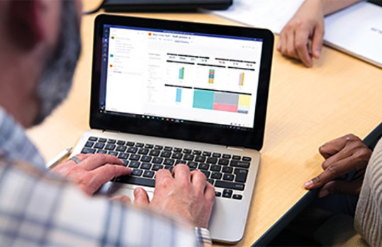 Male teacher sits at table in faculty room, using laptop. Screen shows “Bear Creek Staff > Staff Updates” with staff calendar.