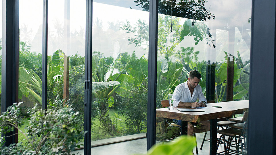 A mature man is sitting at a table in front of a lush garden, writing