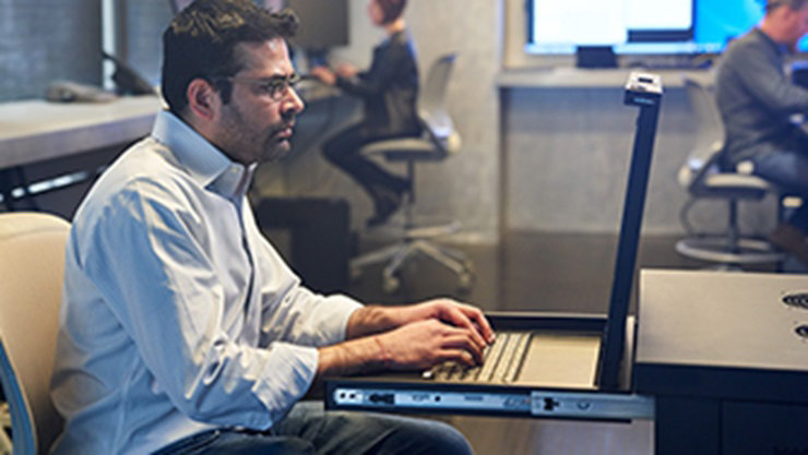 man sitting in an office chair working on a device with other people working in the background