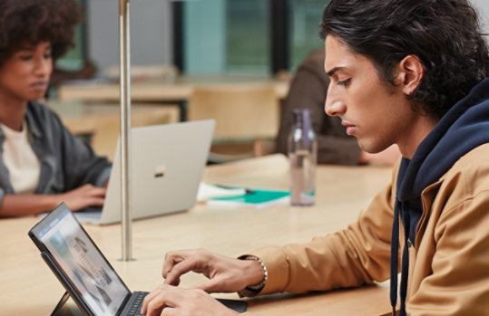 man sitting at a table using a laptop device with another person working on a surface device in the background