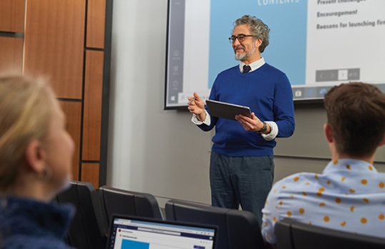 a professor holds a tablet while standing in front of a classroom of students