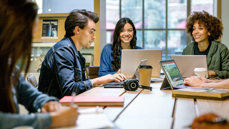 Male and female college students collaborating in group study session in library. Two Surface Pros and two laptops are on table (one Surface Pro screen shows PowerPoint slide; no other screens shown)