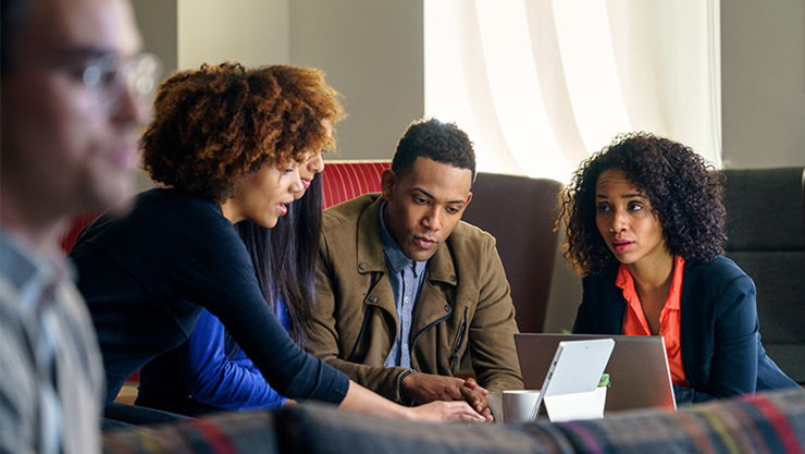 Two male and two female college students in conversation around low table. The woman at left uses a Surface Pro to show something to group.