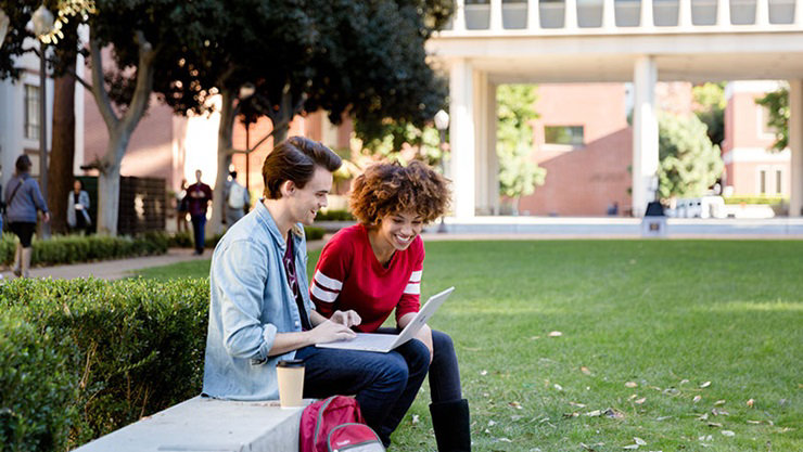 Male and female college students sitting outdoors on campus lawn. The male student is using an open Surface Book (screen not shown) while the female student looks on; both are smiling. Other male and female students walk in background.