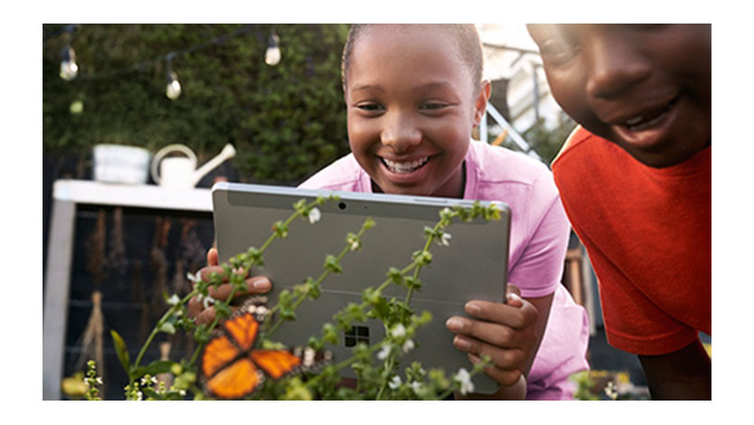 Female K-12 student in a garden holding a Surface Go while taking a picture of a butterfly. Smiling male K-12 student stands beside her.