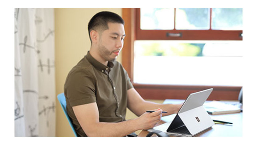 Male teacher sitting at a desk holding a Slim Pen 2 and using a Surface Laptop Studio in studio mode. A Microsoft Arc Mouse sits on the desk.
