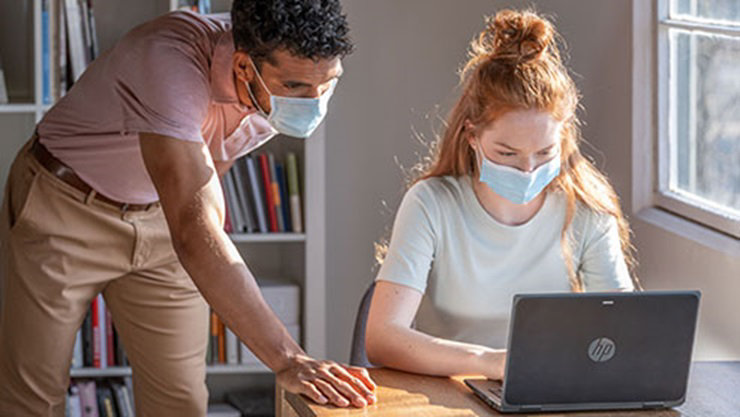 K12 student wearing a mask uses HP Laptop while teacher looks over shoulder in the classroom.