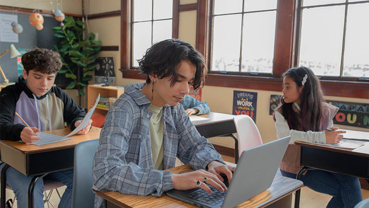 Male and female students sitting at desks in a school classroom. The nearest student is typing on an open laptop and the other students are writing on worksheets.