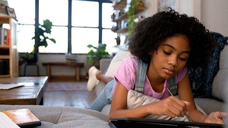 A K-12 student works on a device while laying on a sofa