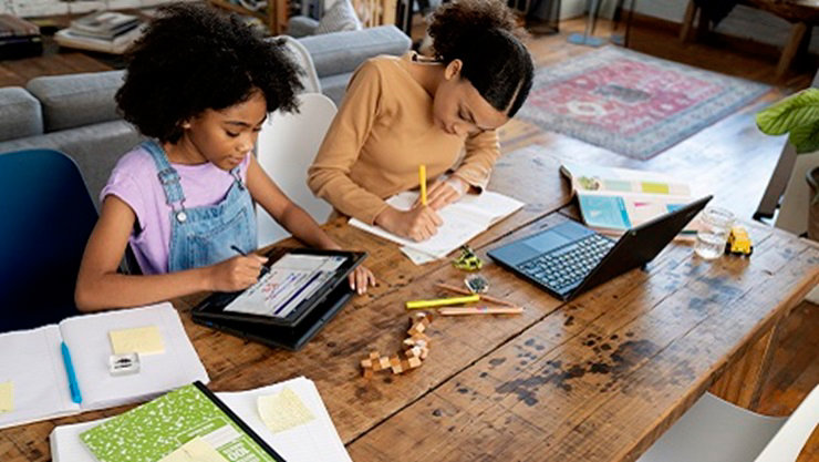 Two K-12 students sit together at a table working on devices and schoolwork