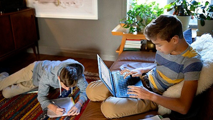 A K-12 student sits on a sofa using Microsoft Teams on a device.  A second K12 student is laying on the floor working on school work.