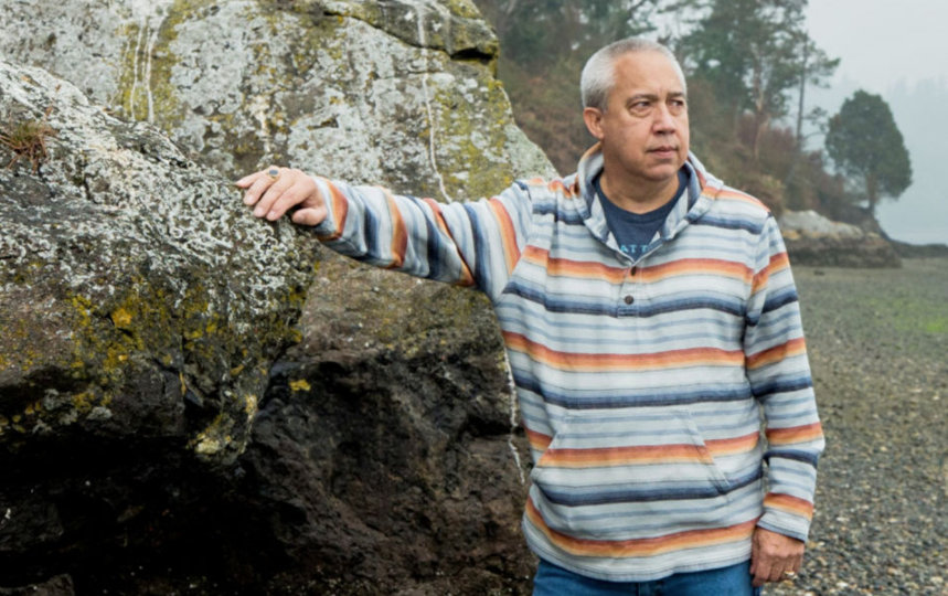 A man stands against a seashore boulder, with his right arm perched onto the boulder, looking out at the water