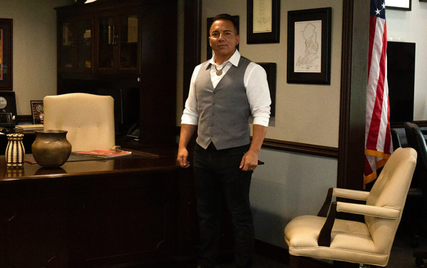 Richard Sneed, principal chief of the Eastern Band of Cherokee Indians, standing in front of the his desk in his office, with a rug of the seal of the EBCI on the floor in front of him.