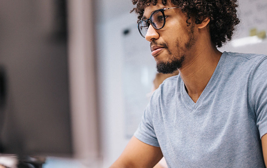 Programer in a blue t-shirt and glasses sits at a row of computer terminals, focusing on the training on his screen