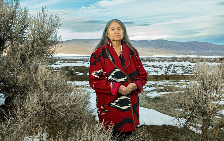 A tribal elder stands against a snowy valley, wearing a red blanket coat looks into the camera