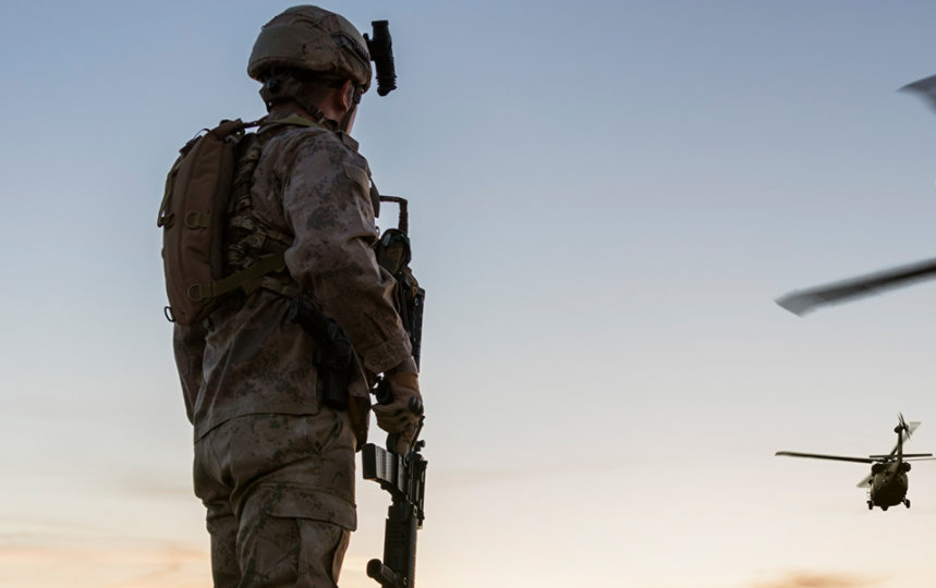 Man standing in combat uniform watching 2 helicopters fly towards the sunrise