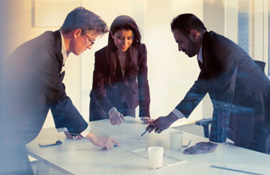 Two business men and a business woman work together, standing over papers on a conference table