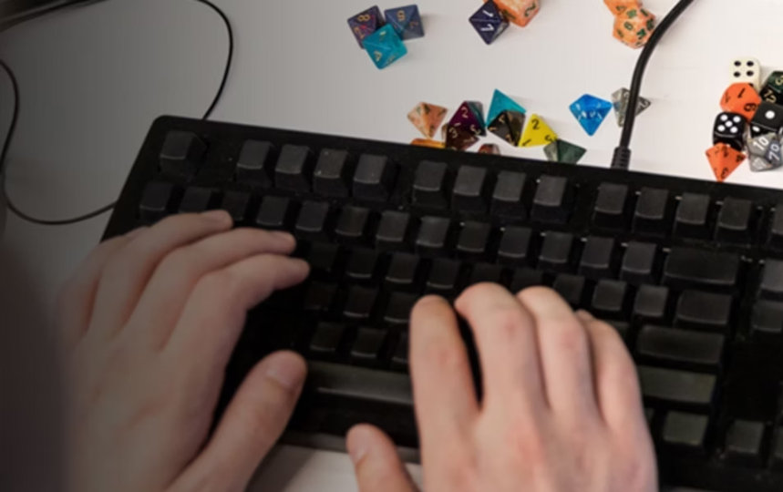 無刻印の黒いメカニカルキーボードでタイピングする手元、白い机の上にカラフルなテーブルトップRPG用ダイスが散らばっている。