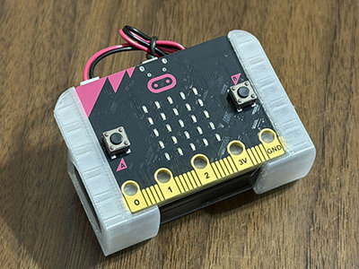 ボタン、LED グリッド、ワイヤー、灰色の側面アタッチメントが付いた micro:bit 基板が木製の面に置かれている。