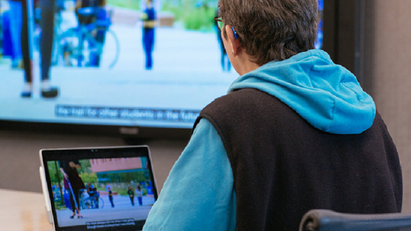 青いパーカーを着た人物が、字幕付きの屋外映像を映すノートPCを使用し、同じ映像が背後の大きなモニターにも表示されている。
