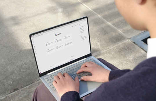 日差しの当たるコンクリートの上で、人物がシルバーのノートパソコンを操作している。画面には Microsoft Copilot のチャット形式の画面が表示され、上部に「Copilot」という文字が見える。