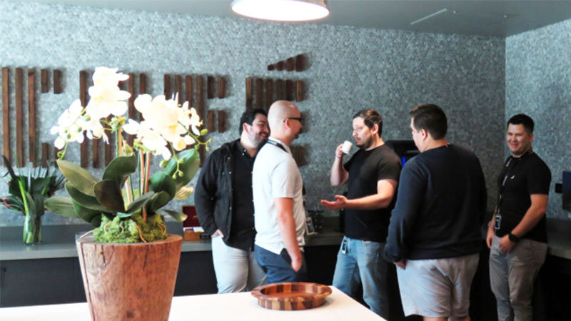 Un jarrón con una flor en primer plano y unas personas hablando al fondo.