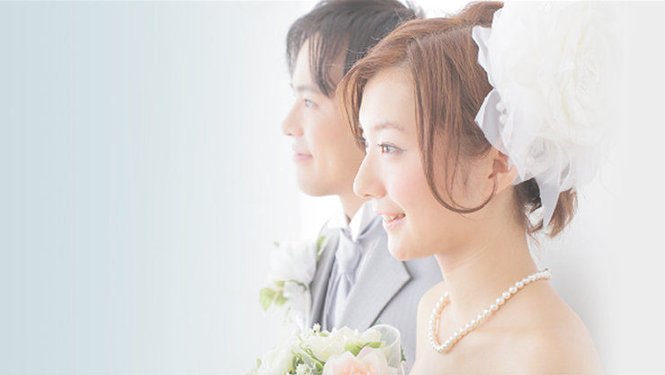  白いドレスとパールネックレスを身につけた笑顔の花嫁が、グレーのスーツを着た新郎の隣に立ち、二人とも前方を見つめている。