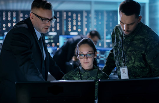 Three national security professionals confer in front of a series of computer screens in a command center
