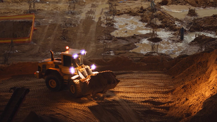 A skid-steer loader moves piles of dirt from the side of a large mound at dusk