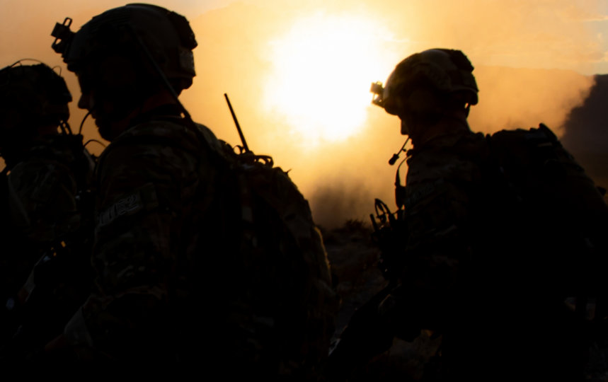 Silhouetted, sunrise view of three soldiers in tactical gear and helmets walking past a grounded helicopter in the background